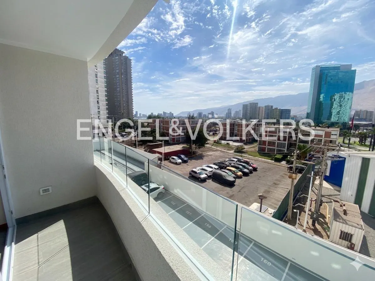 View from a balcony with glass railings overlooking a parking lot, city buildings, and mountains under a blue sky.