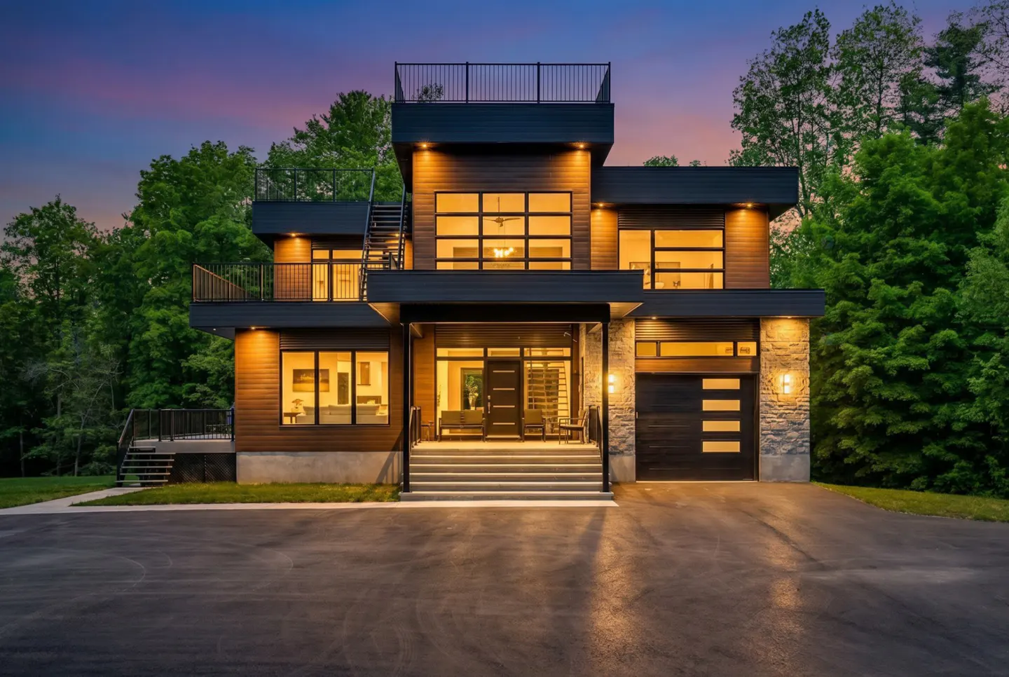 Modern multi-level home with dark wood siding, stone accents, and illuminated windows at dusk, set against a forest backdrop.