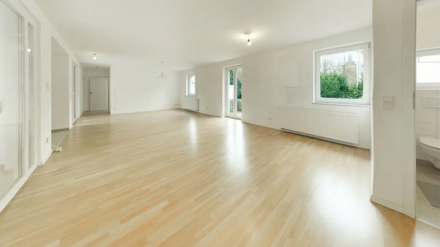 Bright, empty room with light wood floors, white walls, and natural light. A window and glass door offer views of greenery. A bathroom is visible.