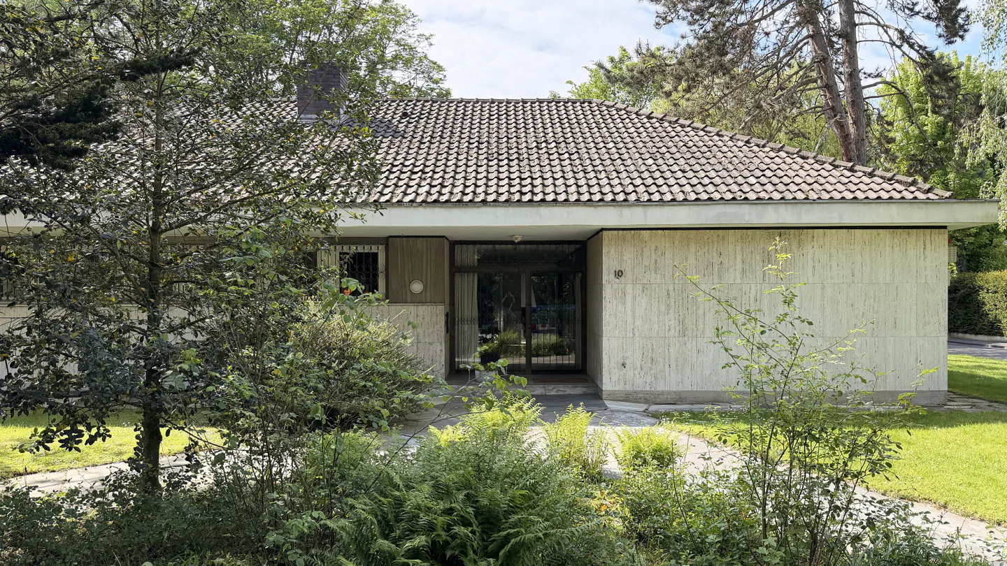 A single-story house with a brown tile roof, stone walls, and glass front door, surrounded by green trees and bushes.