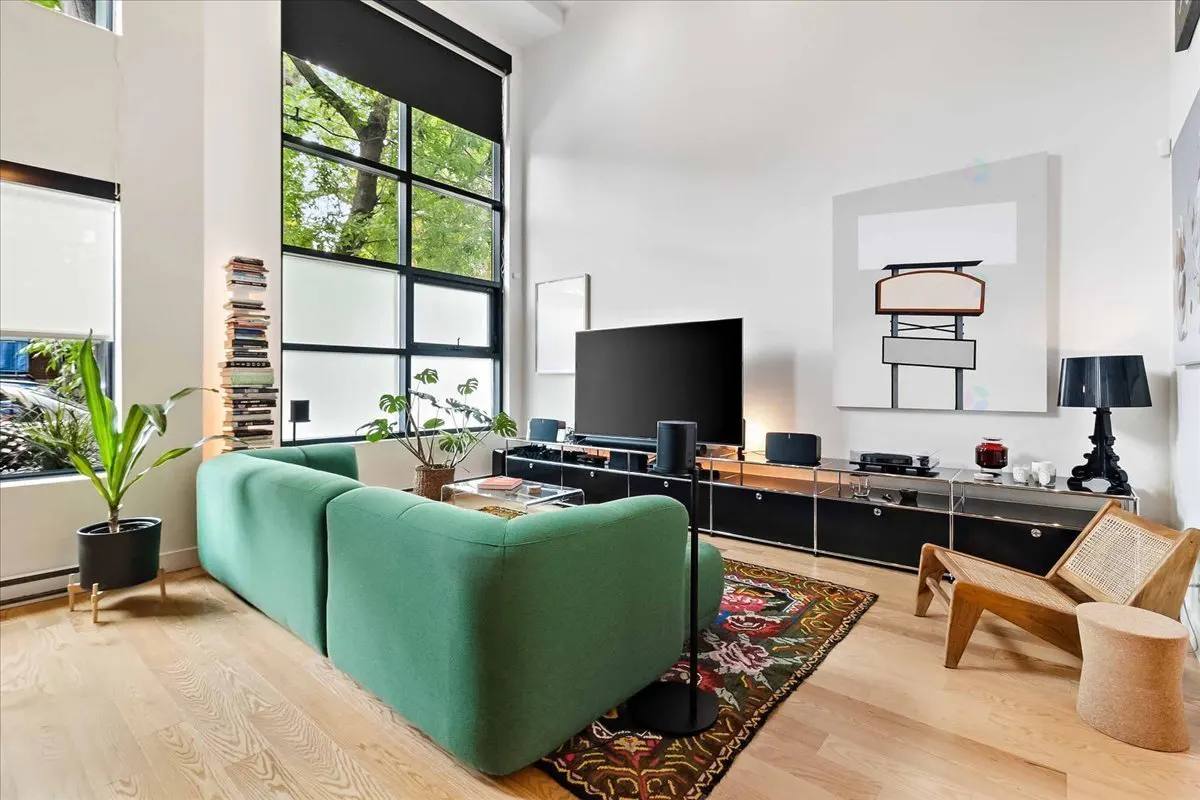 Bright living room with a green sofa, black TV stand, and large windows. Artwork and plants add to the modern decor.