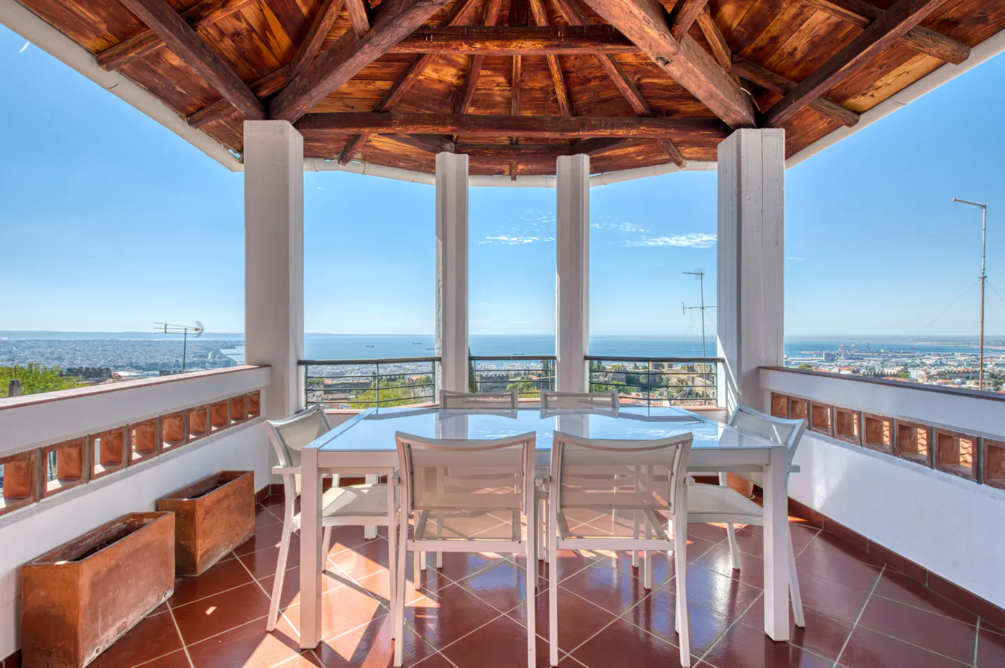 Outdoor dining area with white table and chairs on a tiled patio, overlooking a city and ocean view.