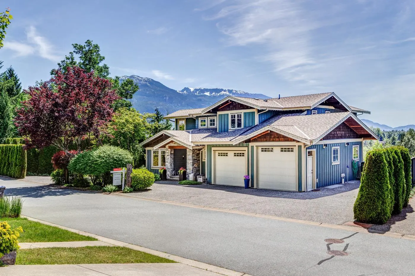 Blue two-story house with a double garage, stone accents, and a mountain backdrop under a clear blue sky.