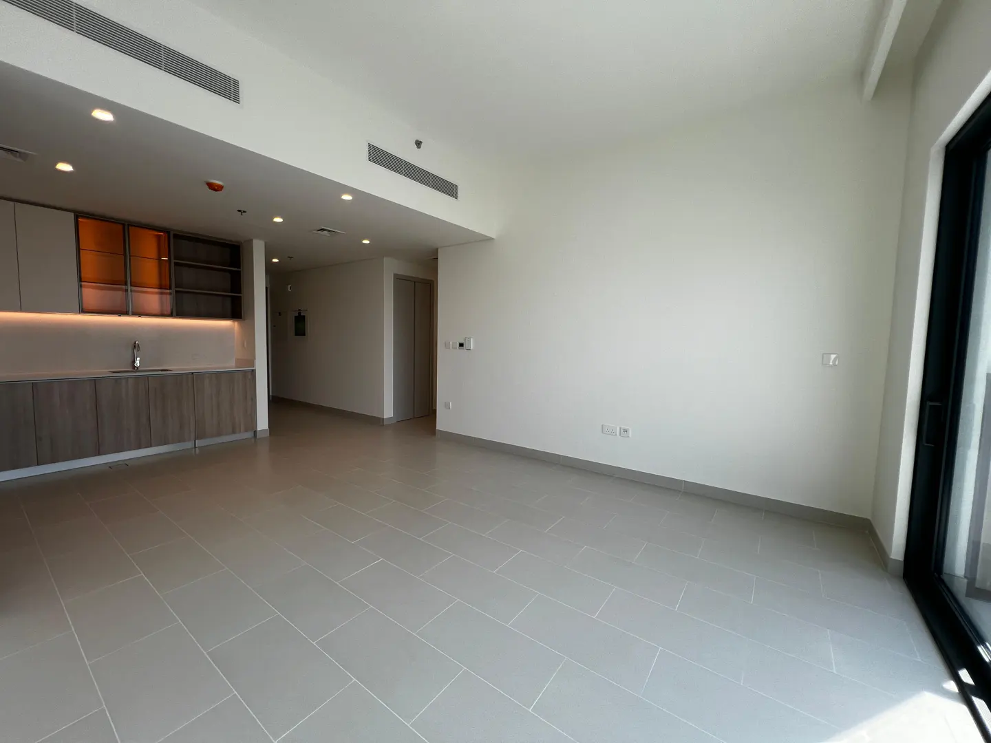 A bright, empty apartment with gray tile floors, white walls, and a modern kitchen with wood cabinets.