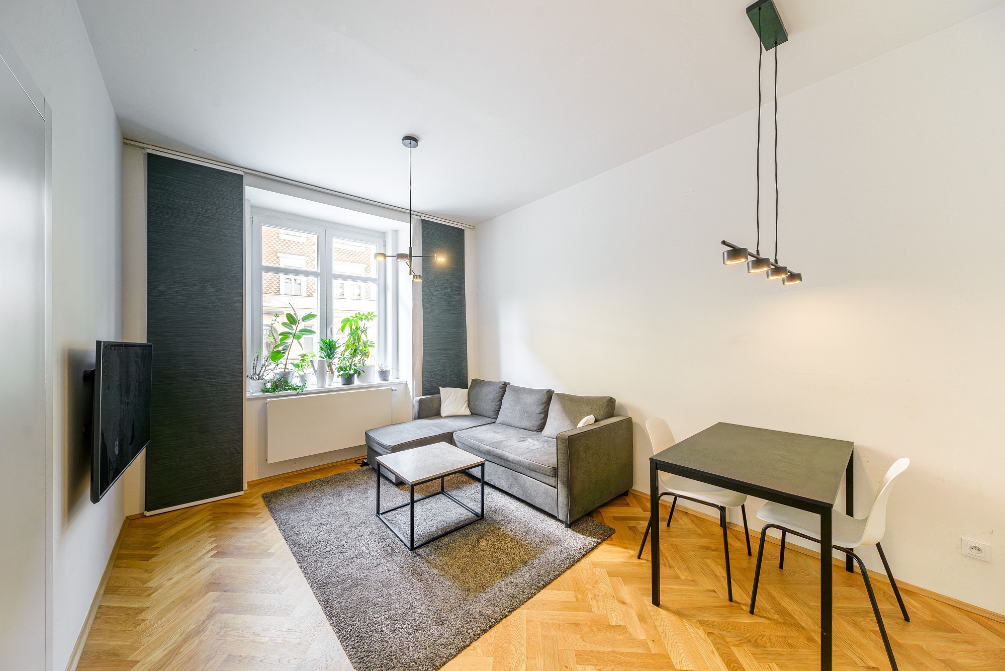 Modern living room with a grey sectional sofa, coffee table, dining set, and plants on a window sill over herringbone floors. [AI-generated text]