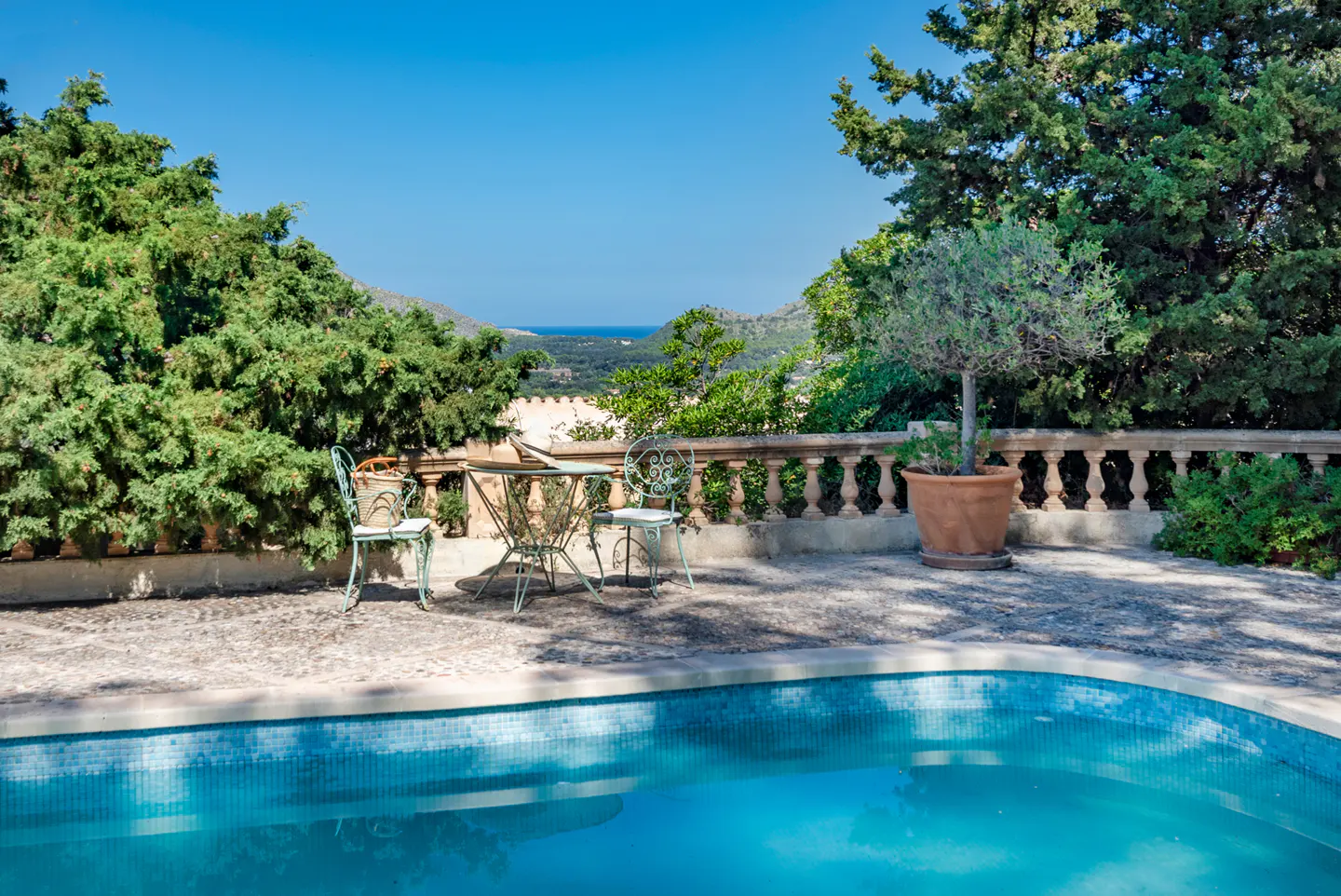Outdoor patio with turquoise pool, table, and chairs. Lush green trees and a blue sky provide a scenic backdrop.