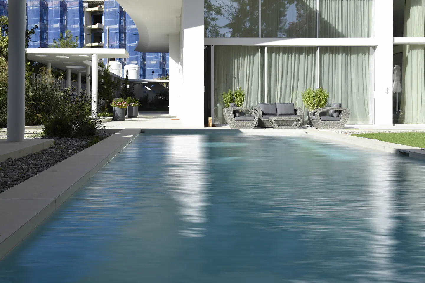 A modern home's pool reflects the white building, outdoor furniture, and greenery.