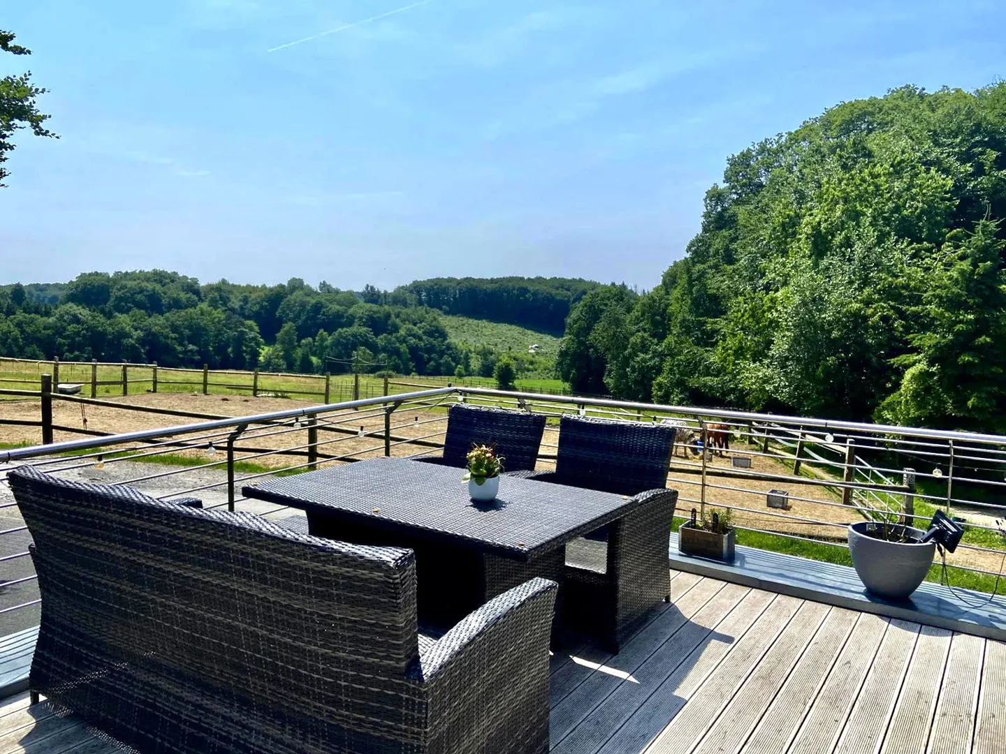 Outdoor patio with a dark wicker table and chairs overlooking a horse paddock and green fields under a blue sky.