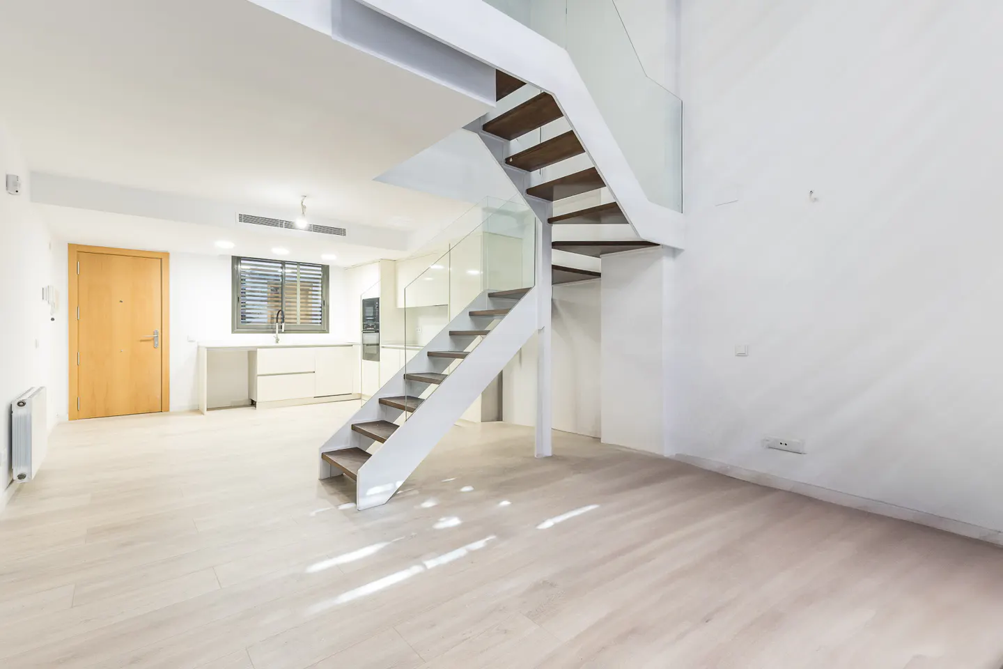Bright, modern interior with white walls, light wood floors, and a staircase with glass railing. A kitchen is visible in the background.