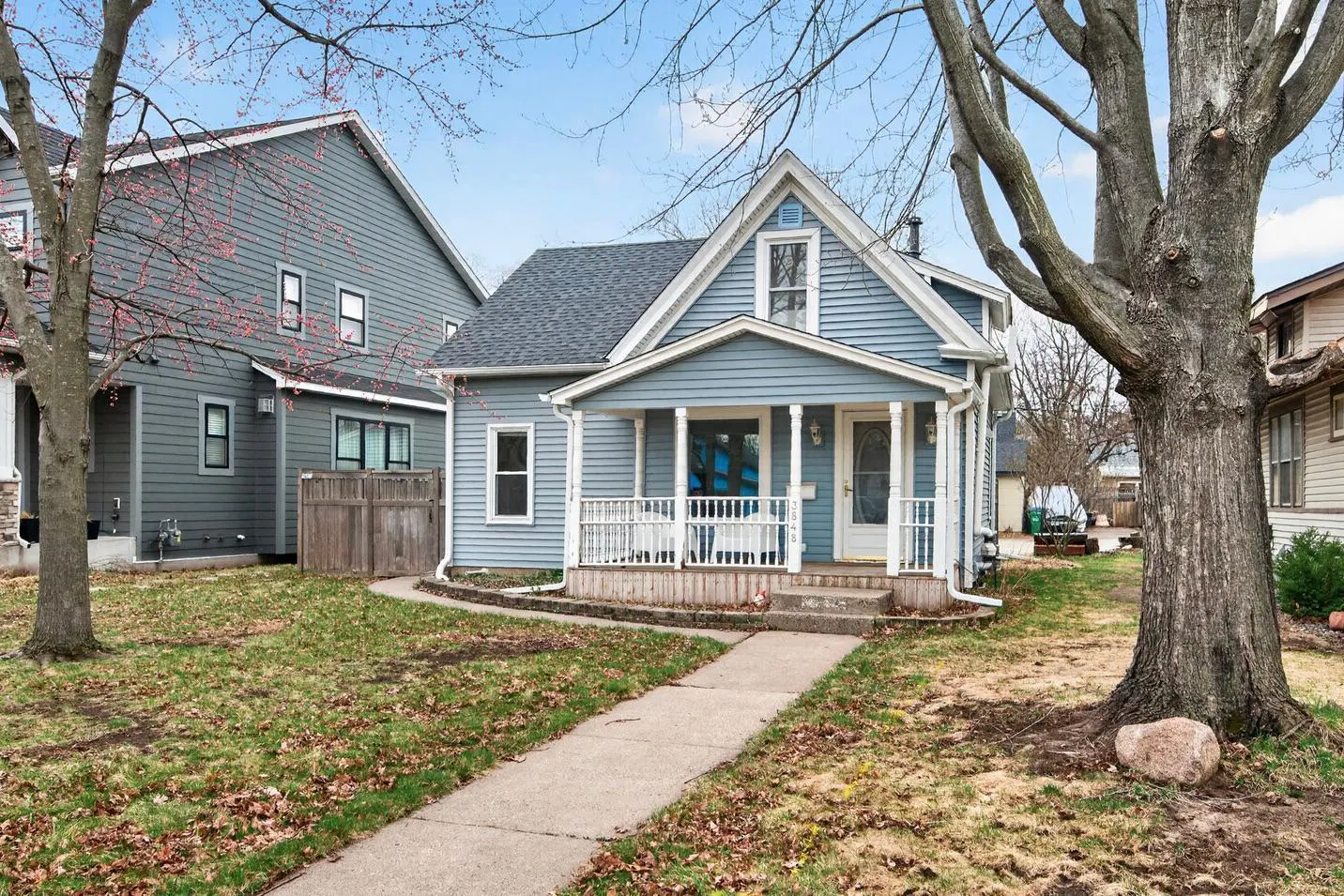 A light blue, two-story house with a white porch and walkway on a lawn with a large tree.