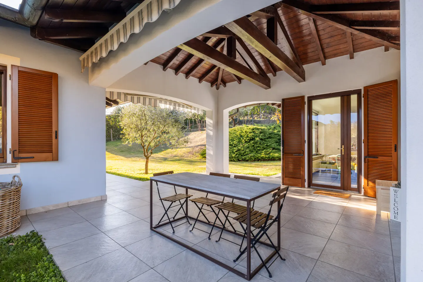 Covered patio with a table and chairs overlooks a green lawn with trees. The patio has brown shutters and a wood-beamed ceiling.