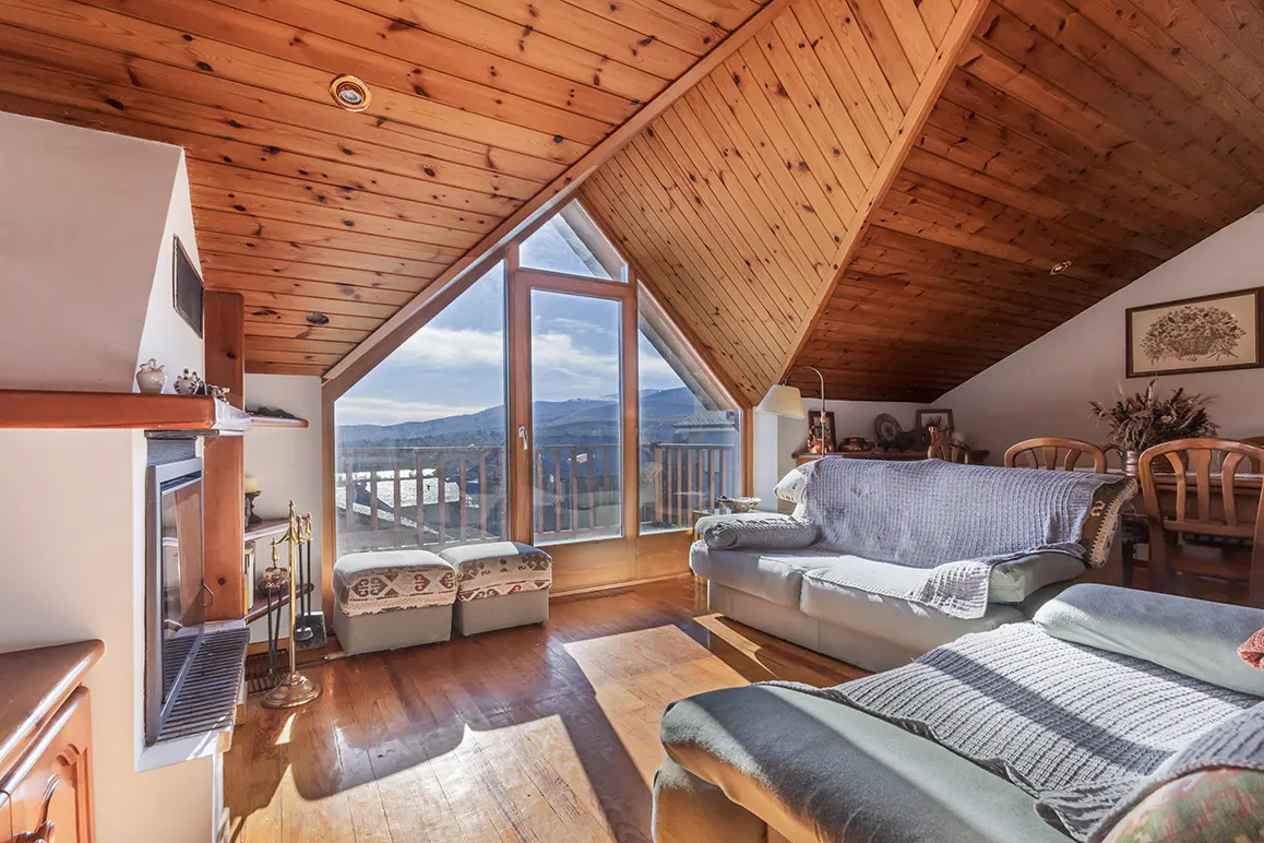 Attic living room with wood-paneled ceiling, fireplace, and large A-frame window with mountain views. Sofas and dining table are visible.