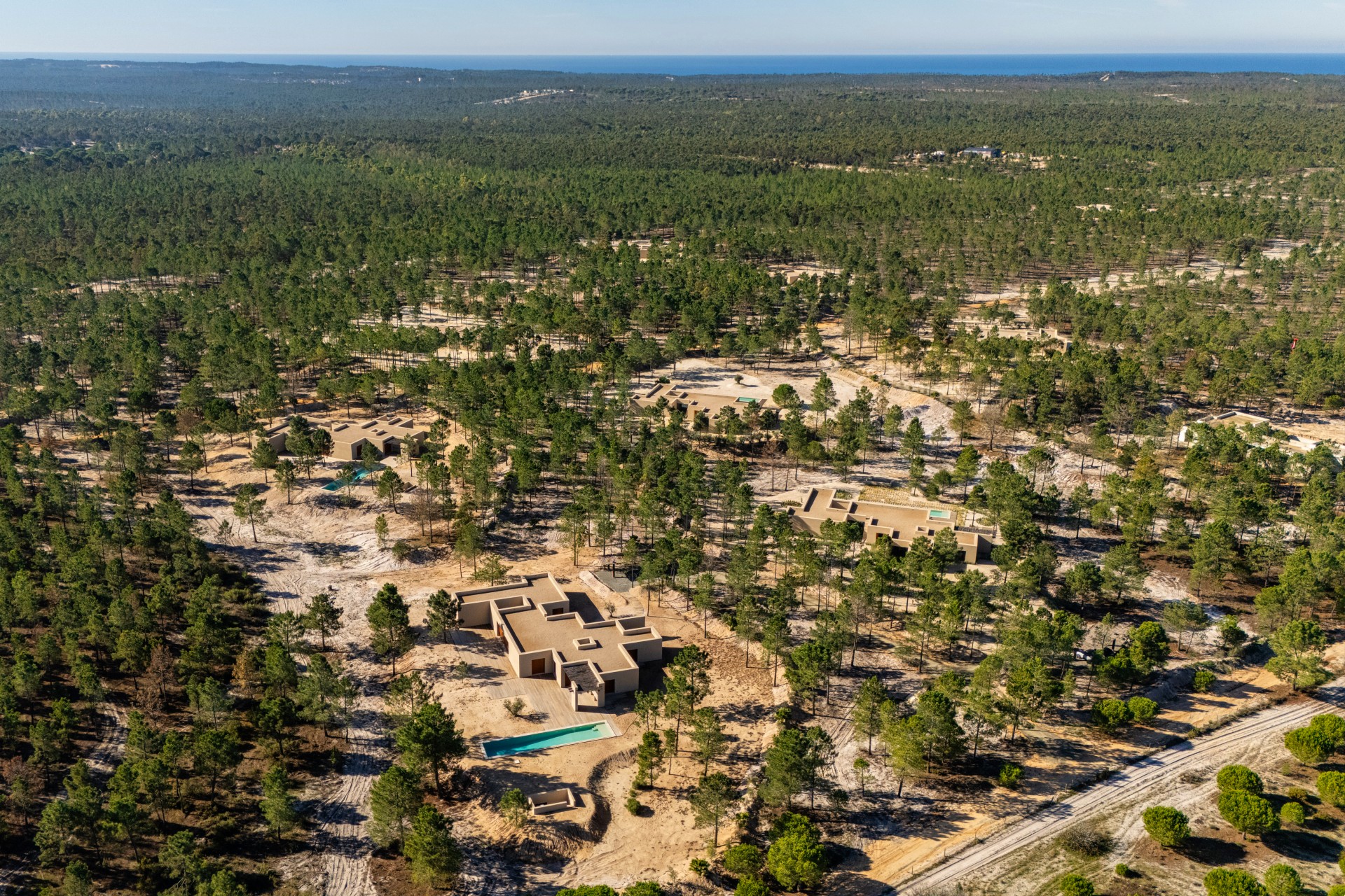 Aerial view of a modern, tan-colored house with a pool nestled in a dense green forest under a clear blue sky.