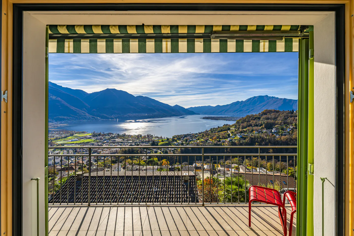 Balcony view of a lake and mountains. A red chair sits on the tiled balcony, with a green and white striped awning above.