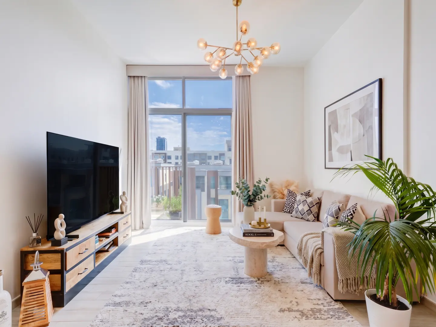 Bright living room with beige sectional sofa, patterned rug, and large TV. Balcony visible through floor-to-ceiling windows.