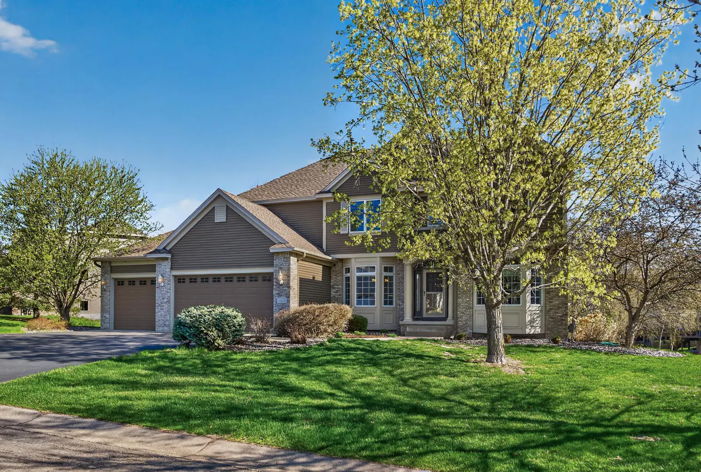 Two-story brown house with a three-car garage, stone accents, and a green lawn under a blue sky.