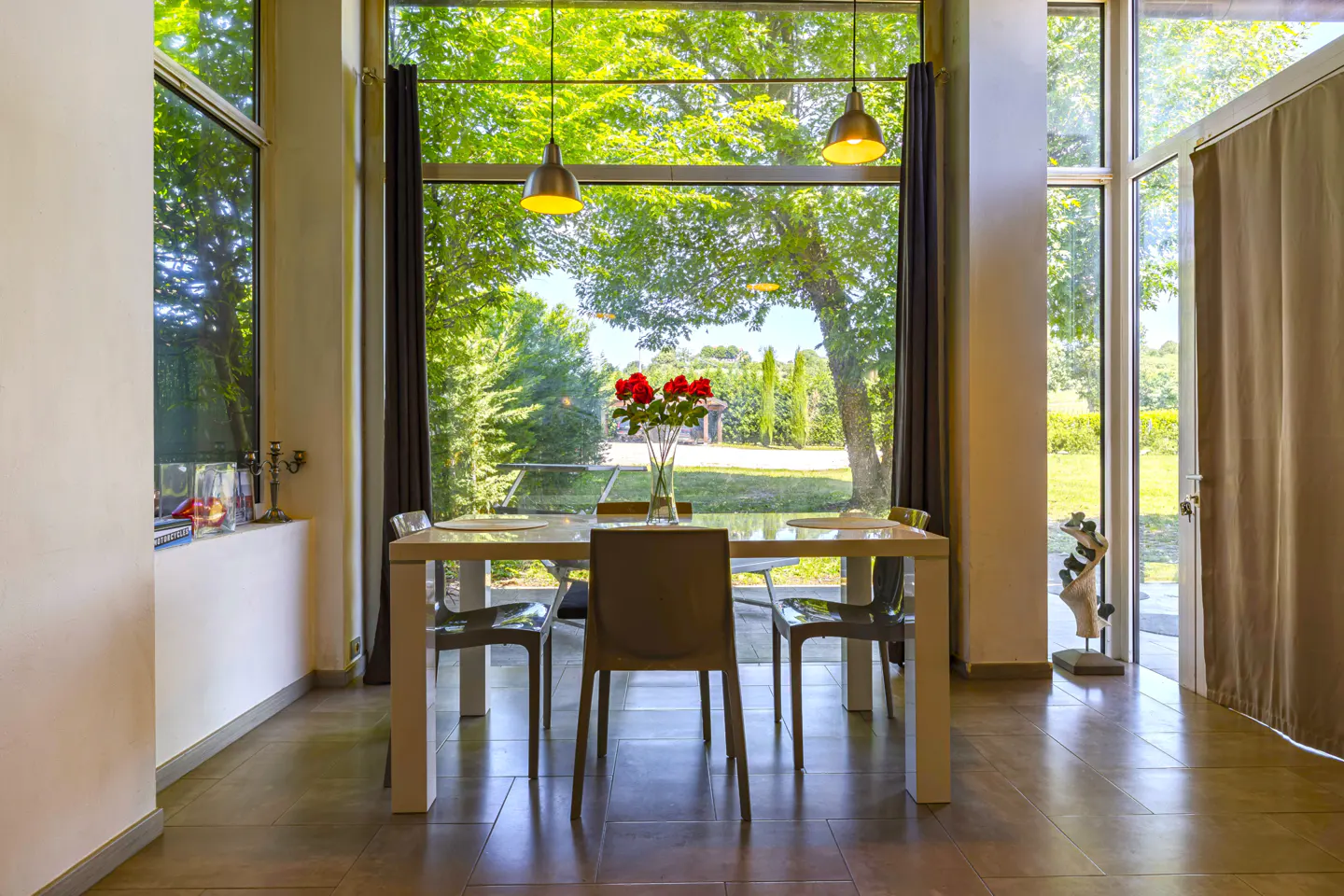Dining room with a white table, chairs, and a vase of red roses. Large windows overlook a green lawn and trees.