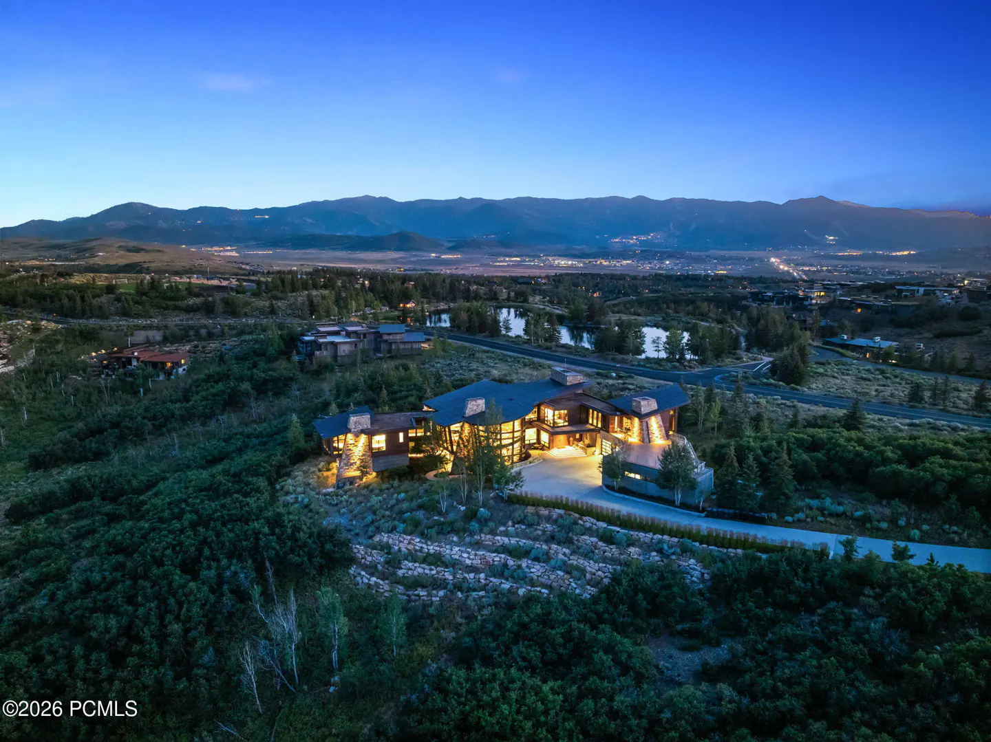 Aerial view of a large, modern home with dark roof, surrounded by green trees and mountains at dusk.