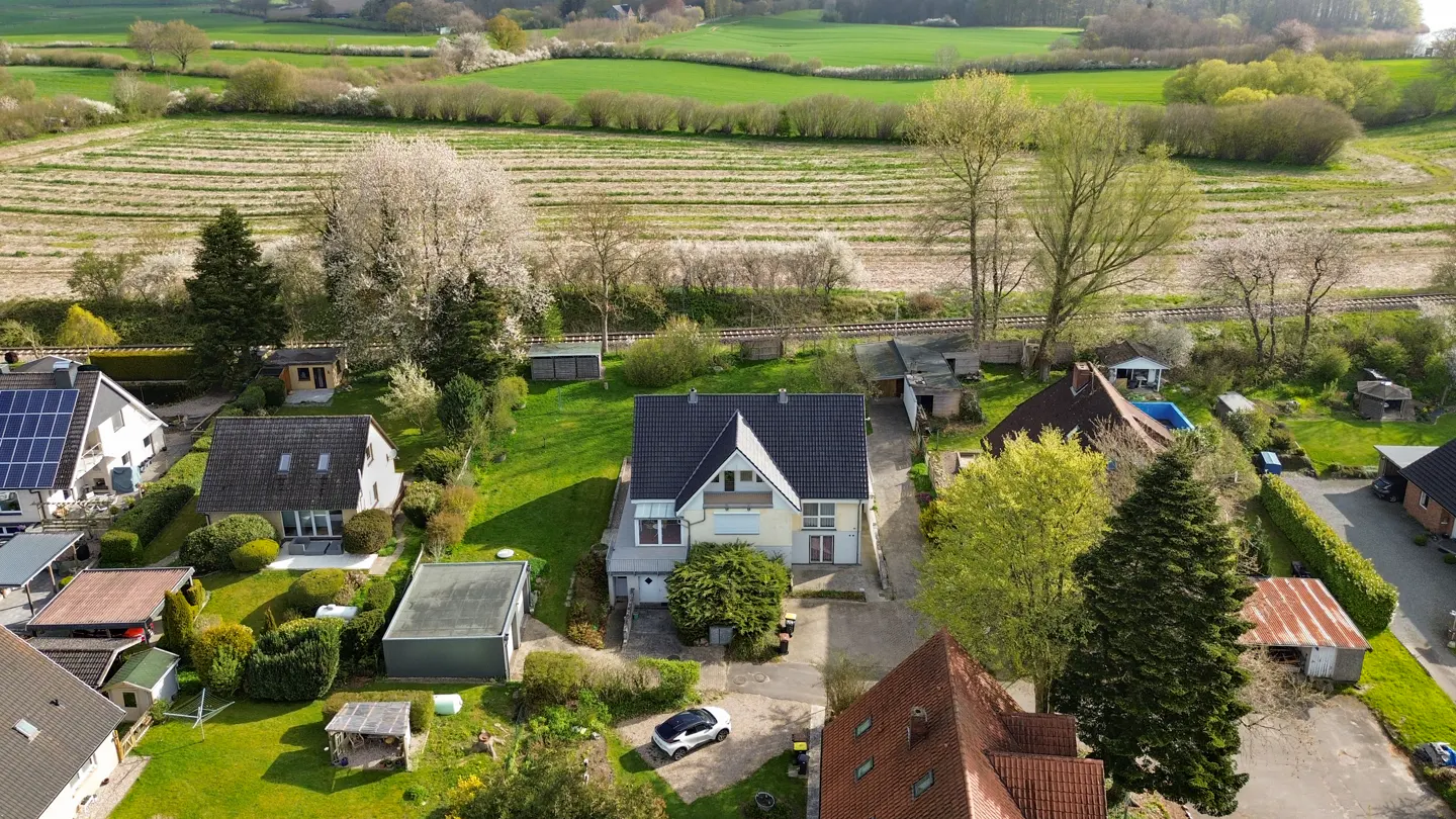 Aerial view of a light yellow two-story house with a dark gray roof, surrounded by green lawns, trees, and other houses.