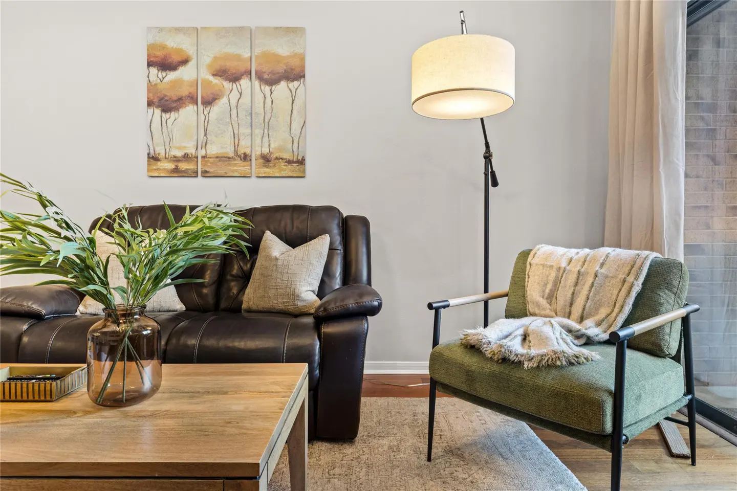 Living room with a brown leather sofa, green chair with throw, floor lamp, and tree art.