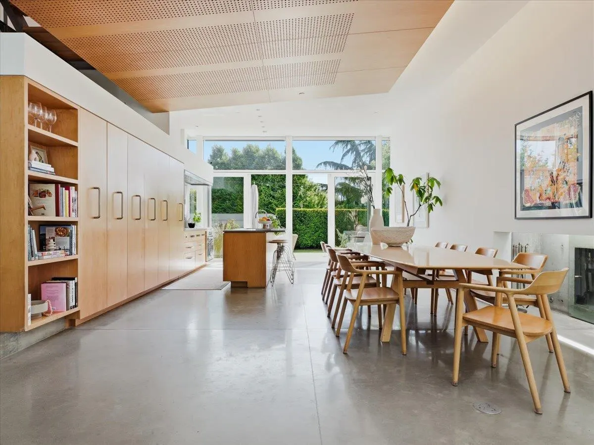 Bright, modern dining room with a long wooden table and chairs. Large windows overlook a green garden. Built-in cabinets line the wall.
