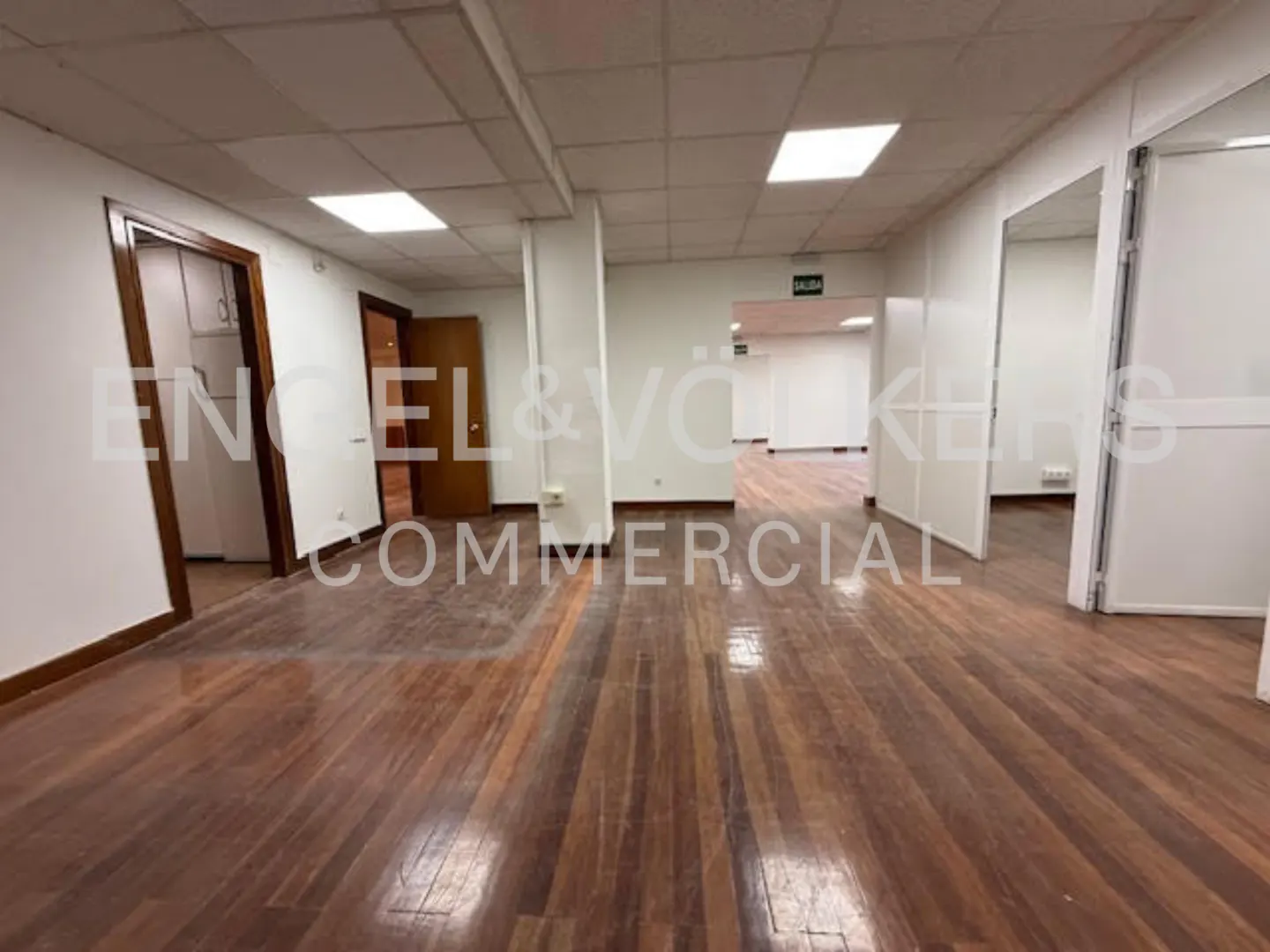 Empty office space with hardwood floors, white walls, and recessed lighting. Doors and hallways lead to other rooms.