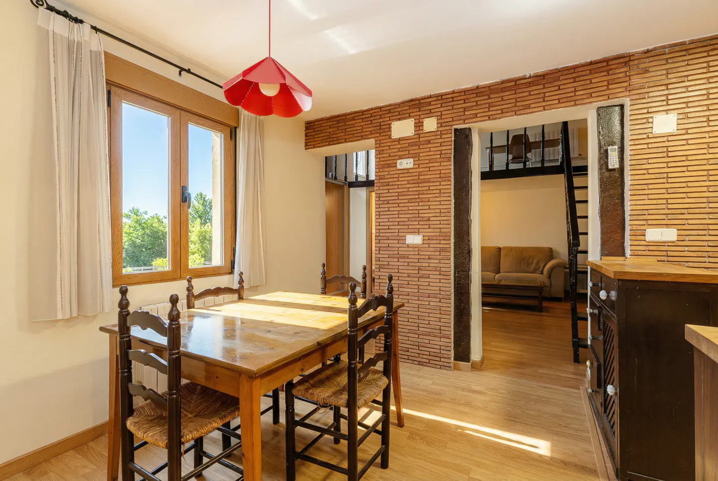 A dining area with a wooden table and chairs, a red pendant light, and a view into a living room with a couch and stairs.