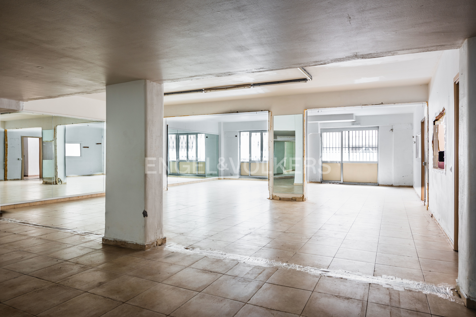 A large, empty room with tile floors and white walls. Mirrors line the walls, reflecting the space. Natural light streams in from windows.