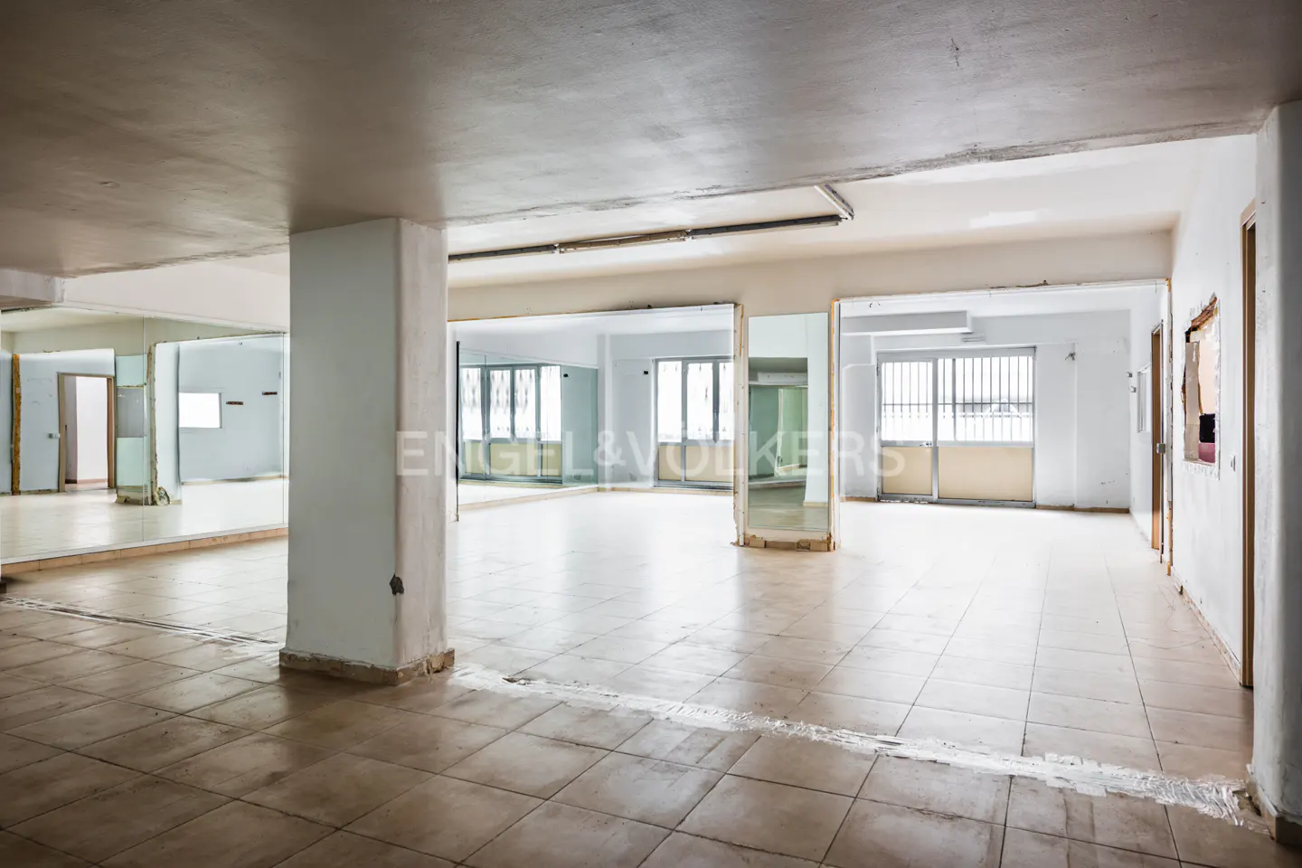 A large, empty room with tile floors and white walls. Mirrors line the walls, reflecting the space. Natural light streams in from windows.