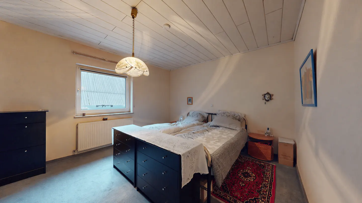 Bedroom with twin beds, black dressers, and a red rug. A window provides natural light, and a decorative lamp hangs from the white, paneled ceiling.