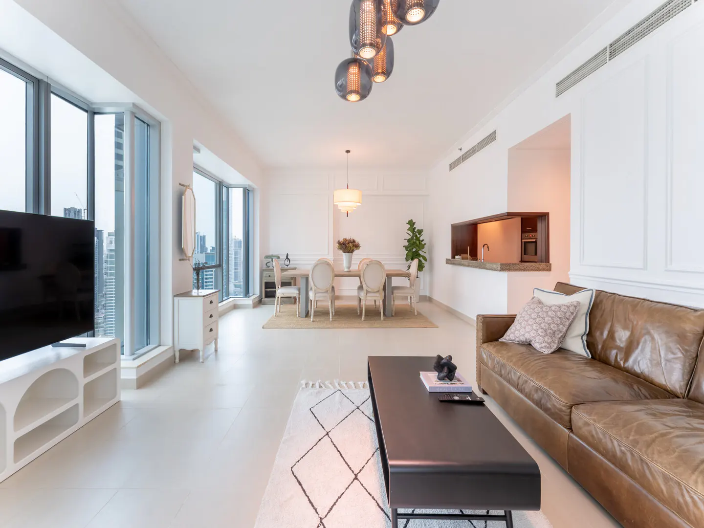 Bright, modern living room with a brown leather sofa, black coffee table, dining table, and large windows with a city view.