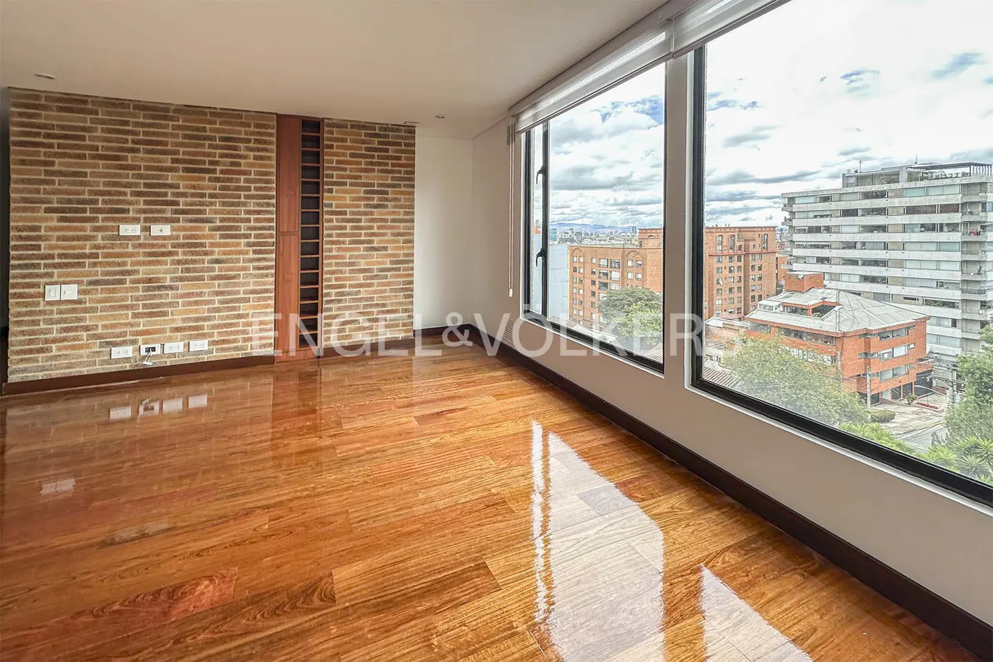 Empty room with polished wood floors, a brick accent wall, and large windows overlooking a city skyline.
