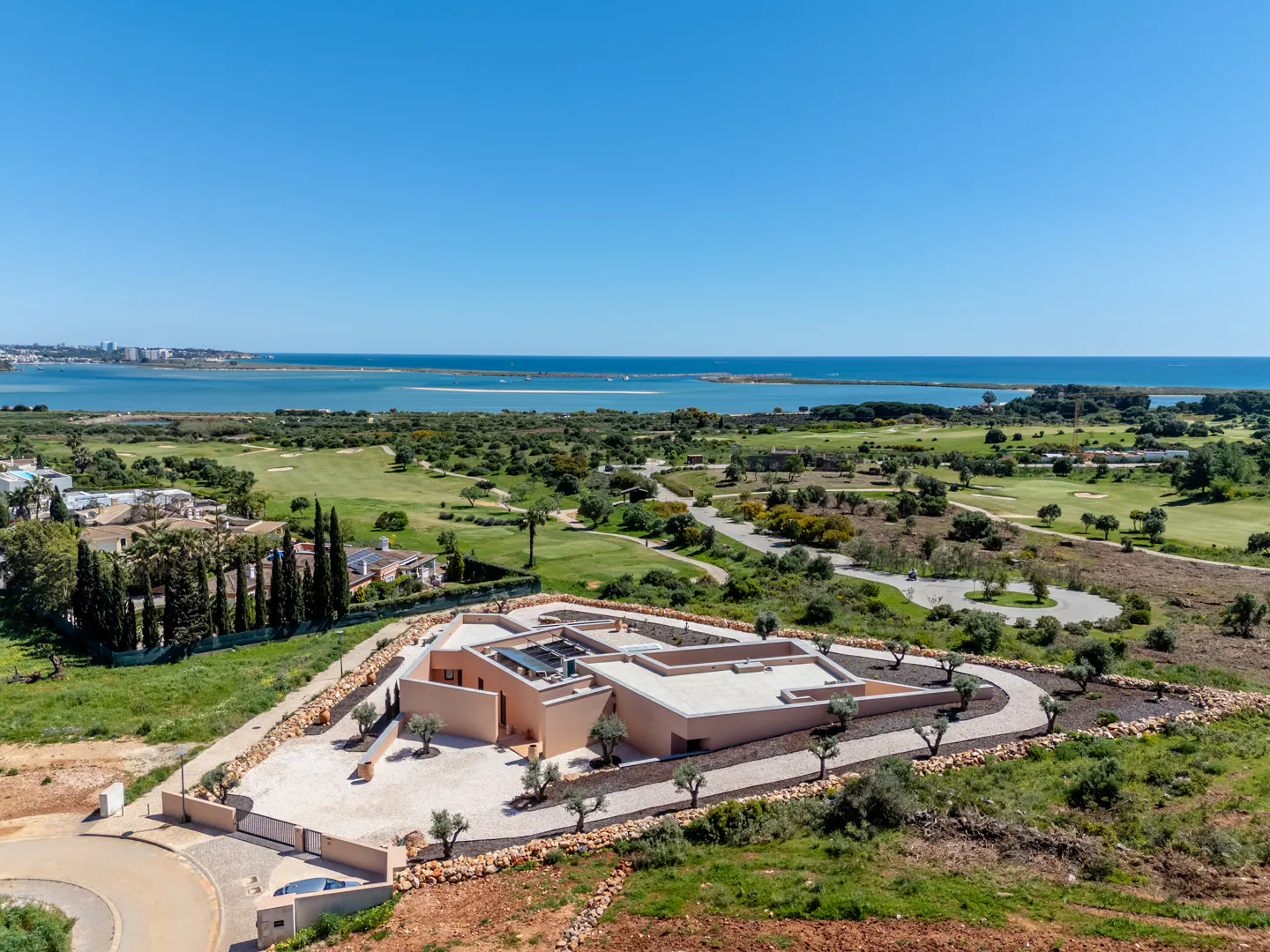 Aerial view of a modern, beige house with a flat roof, surrounded by a golf course, trees, and the ocean under a clear blue sky.