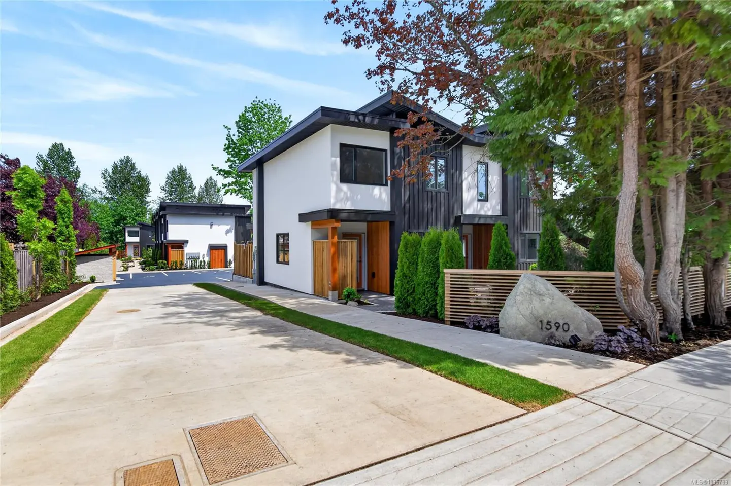 Modern townhouses with white and dark wood exteriors, green landscaping, and a concrete driveway under a blue sky.