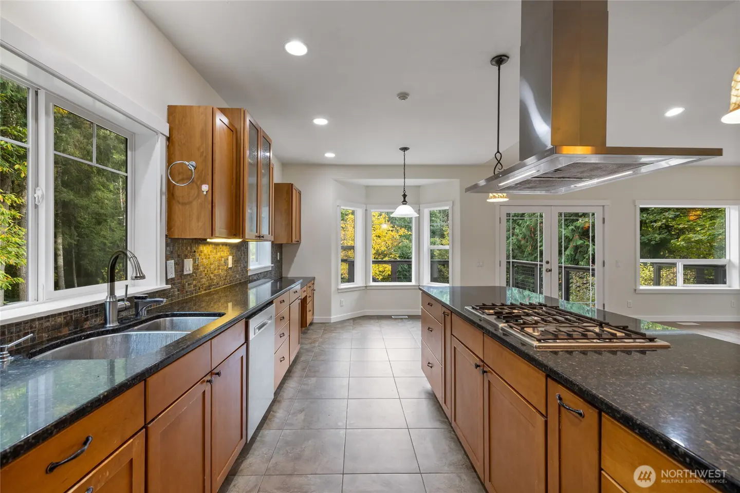 Bright kitchen with wood cabinets, black granite countertops, and stainless steel appliances. Large windows overlook trees.