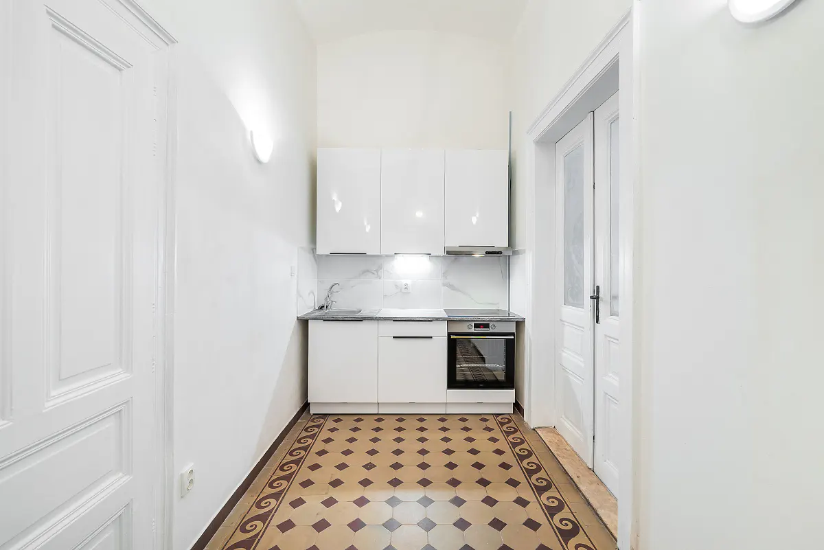Small, bright kitchen with white cabinets, marble backsplash, and patterned tile floor. White walls and doors frame the space.