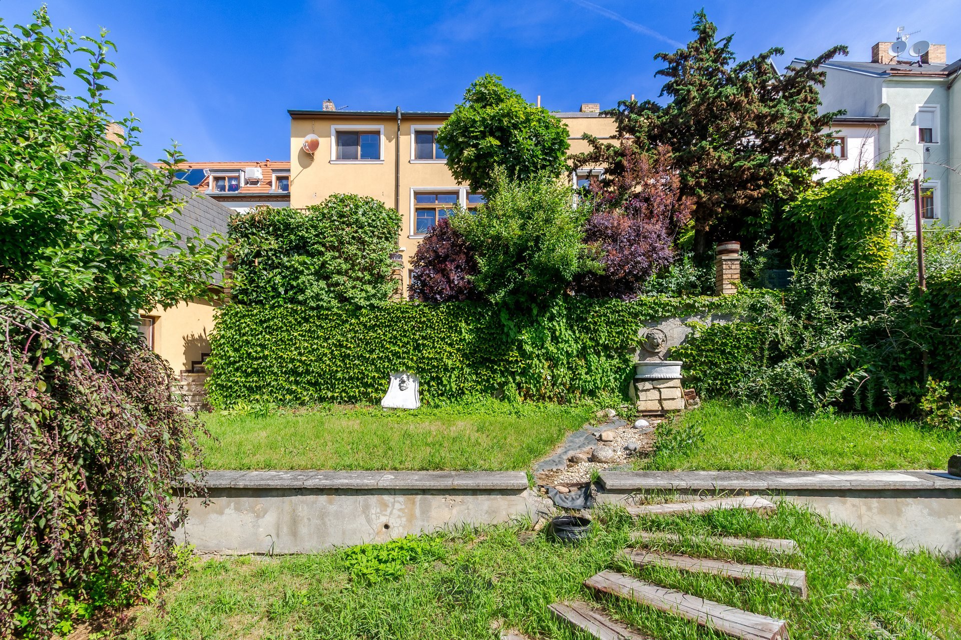 A lush backyard with a stone retaining wall, wooden steps, a small fountain, and a yellow building under a blue sky.
