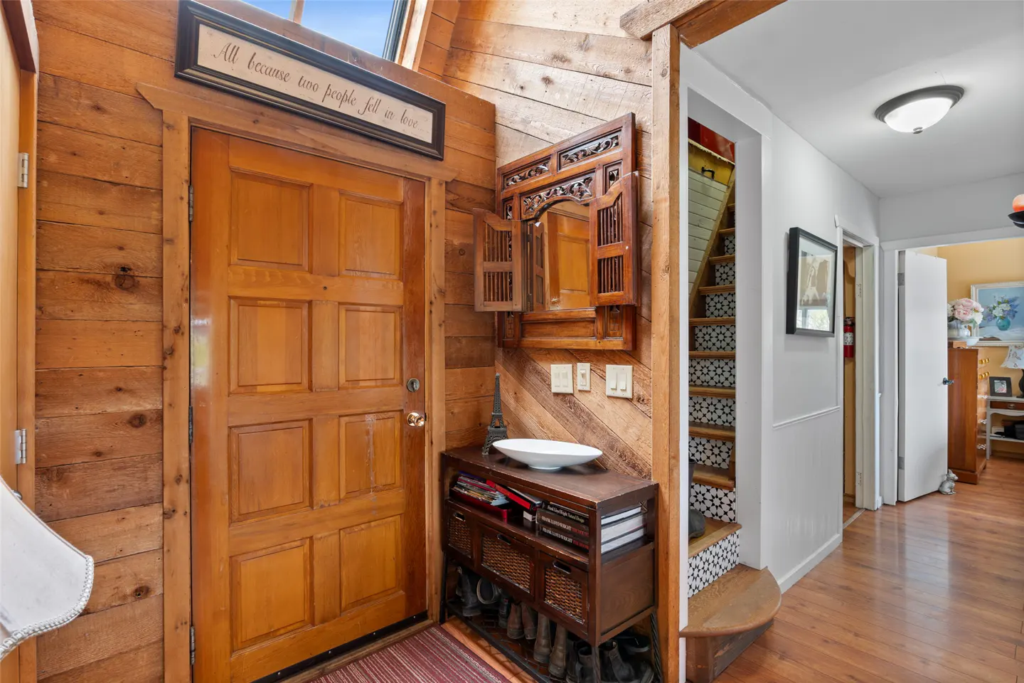 A wood-paneled entryway with a front door, a decorative mirror, and a shoe rack. Stairs with patterned risers lead to the upper level.