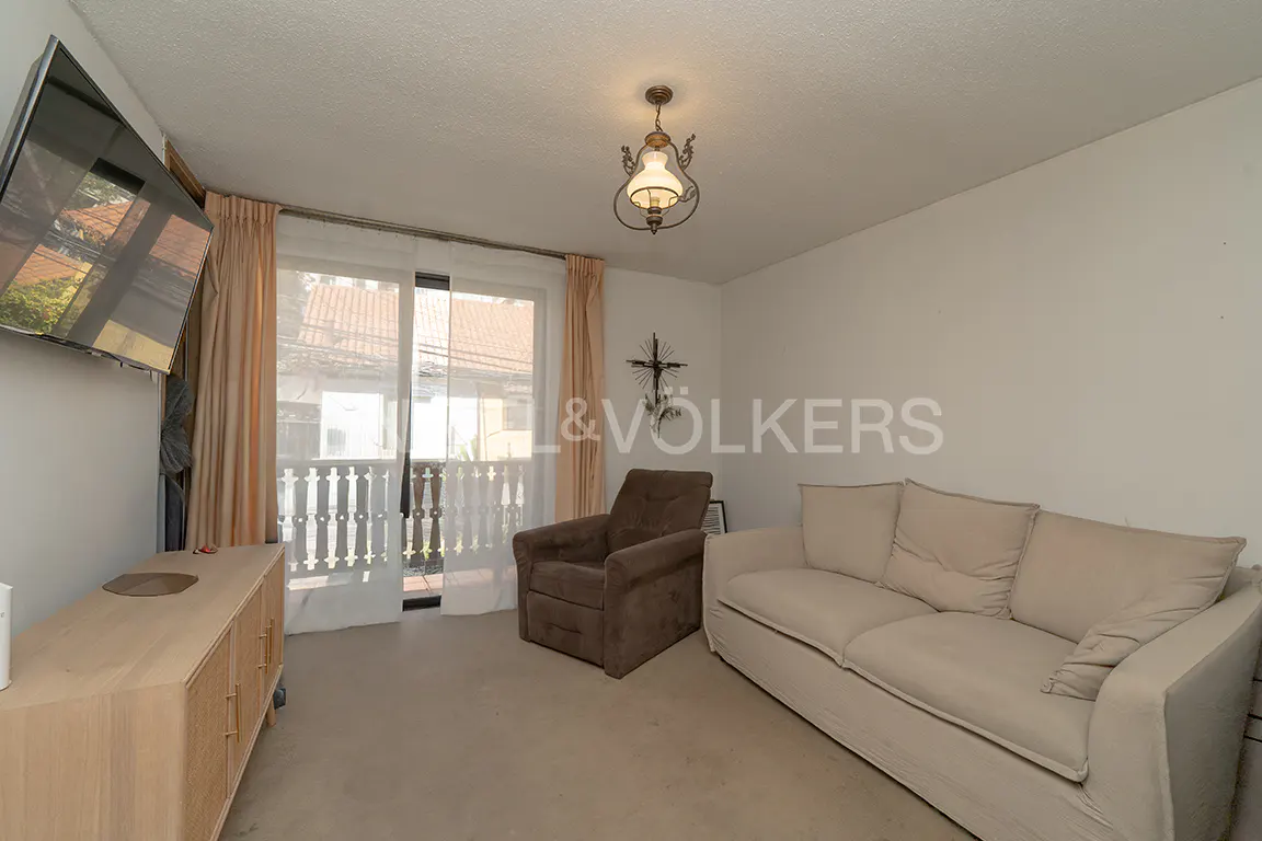 Living room with a beige sofa, brown armchair, TV, and light wood cabinet. Balcony doors with white railings and beige curtains. Overhead light fixture.