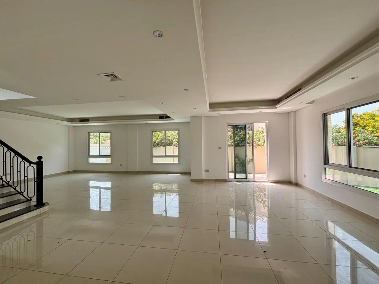 Bright, empty living room with white walls, tile floors, and large windows. A black staircase is visible on the left.