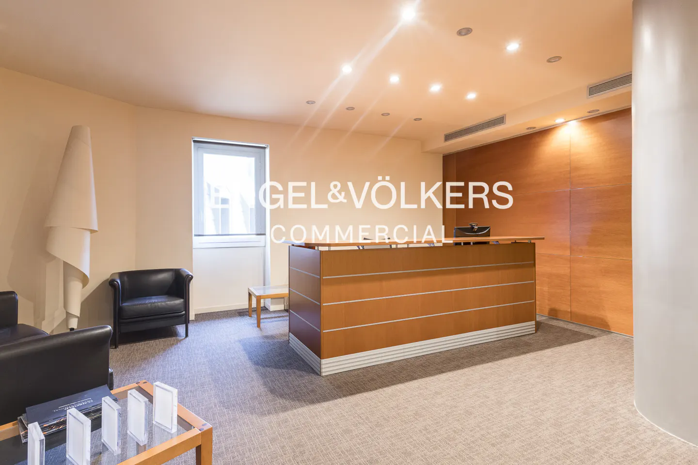 Office reception area with a wood reception desk, black leather chairs, and a glass coffee table. The Engel & Völkers logo is visible.