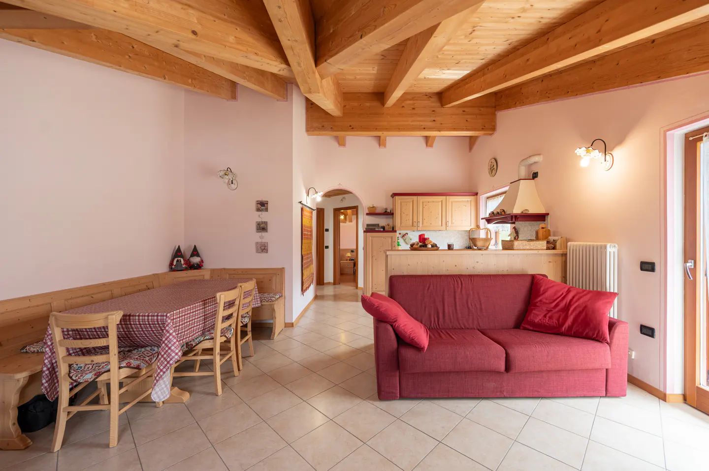 Open-concept living space with a red sofa, wooden dining set, and kitchen area under a wooden beamed ceiling.
