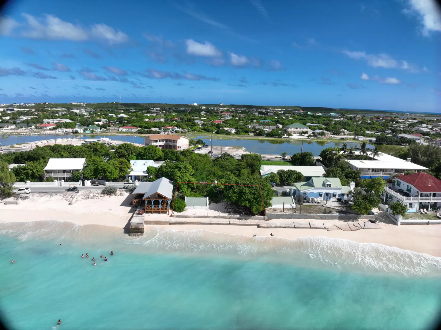 Aerial view of a beach with turquoise water, white sand, and houses along the shore under a blue sky with scattered clouds.