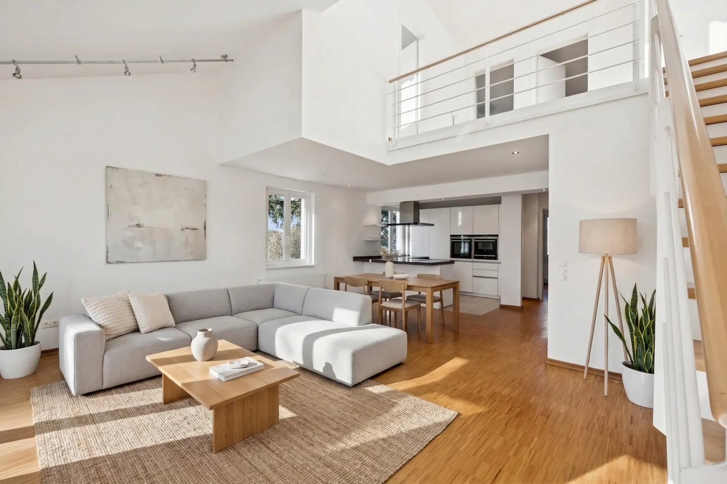 Bright, open-concept living space with a gray sectional sofa, wood floors, and a modern kitchen in the background. Stairs lead to a loft area.