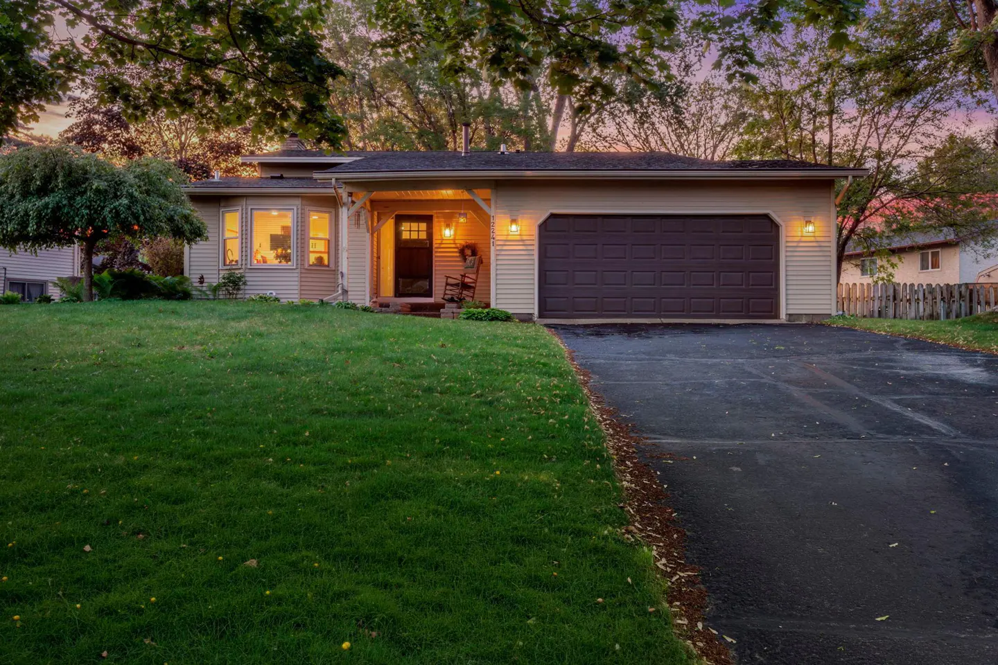 Tan ranch-style house with a dark brown garage door and front door. A black driveway leads to the garage. Green lawn and trees surround the house.