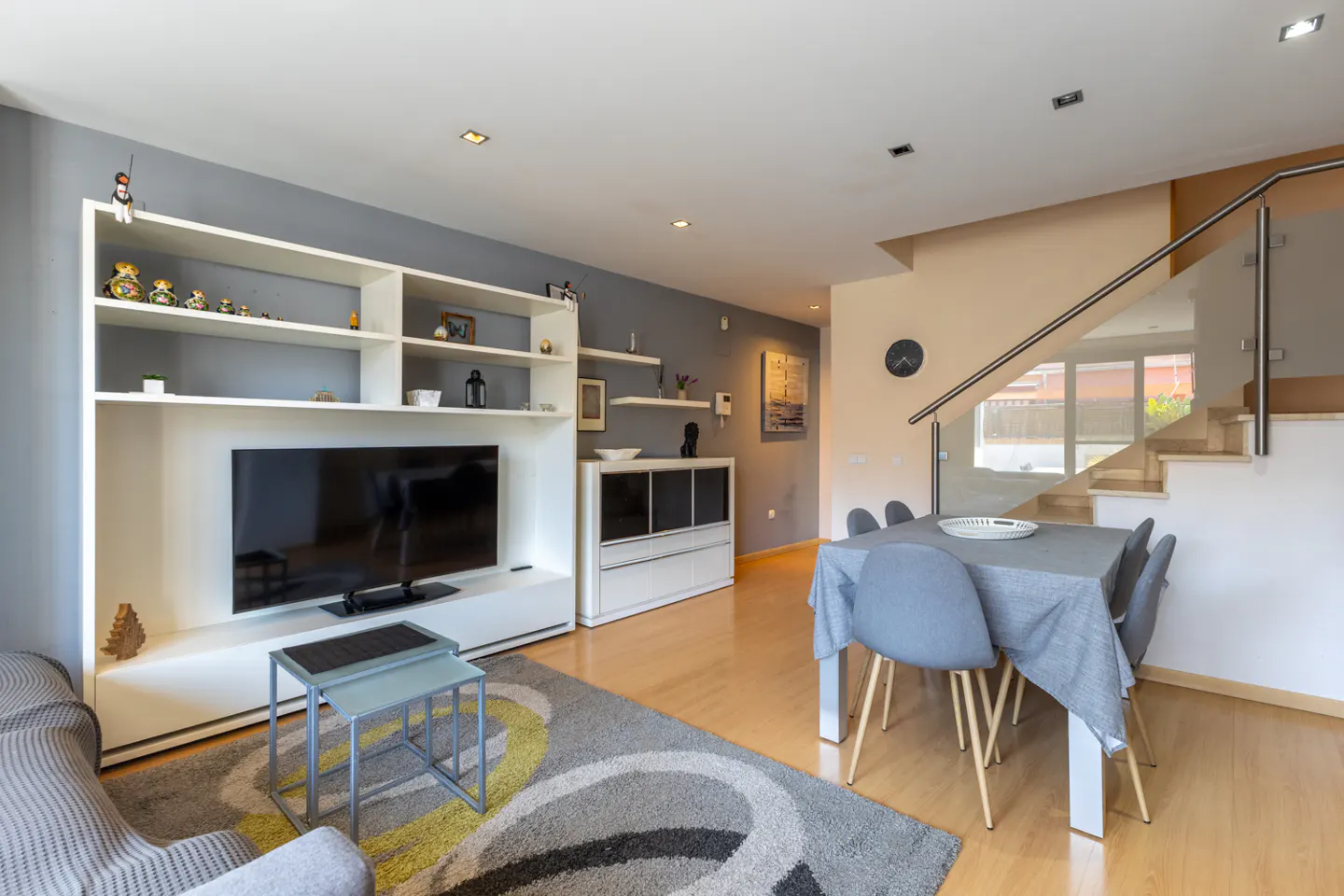 Living room with a TV, white shelves, gray walls, and a dining table with gray chairs. Stairs with glass railing in the background.