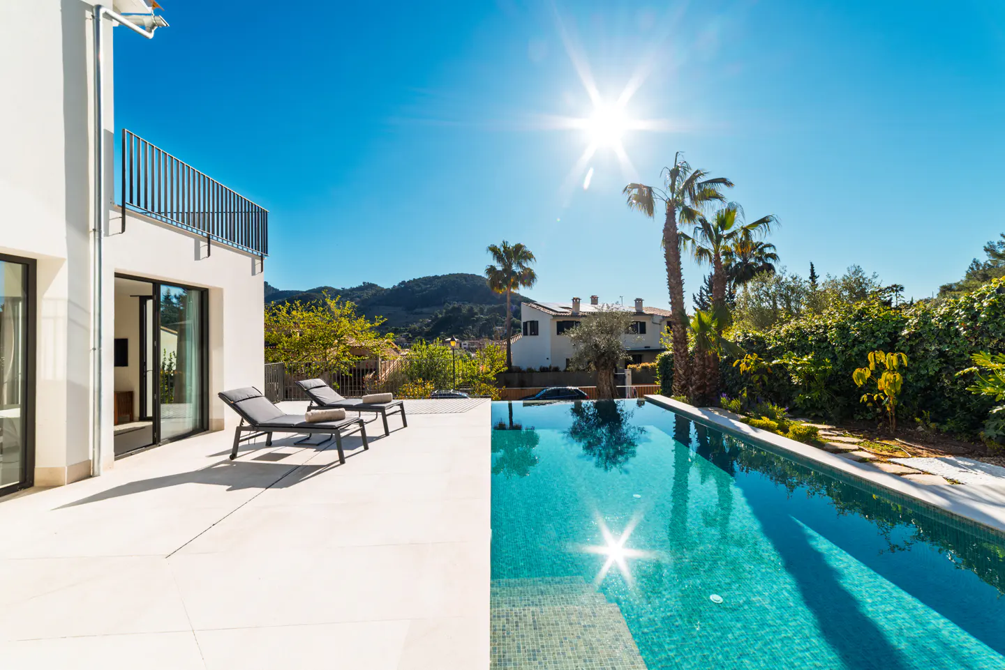 Bright sunny view of a modern white house with a pool, two lounge chairs, and palm trees against a blue sky.