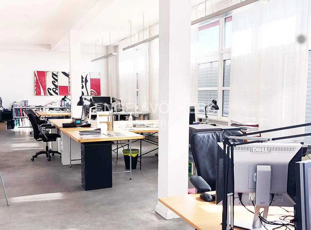 Bright, modern open-plan office with light wood desks, black chairs, concrete floors, and Coca-Cola wall art.