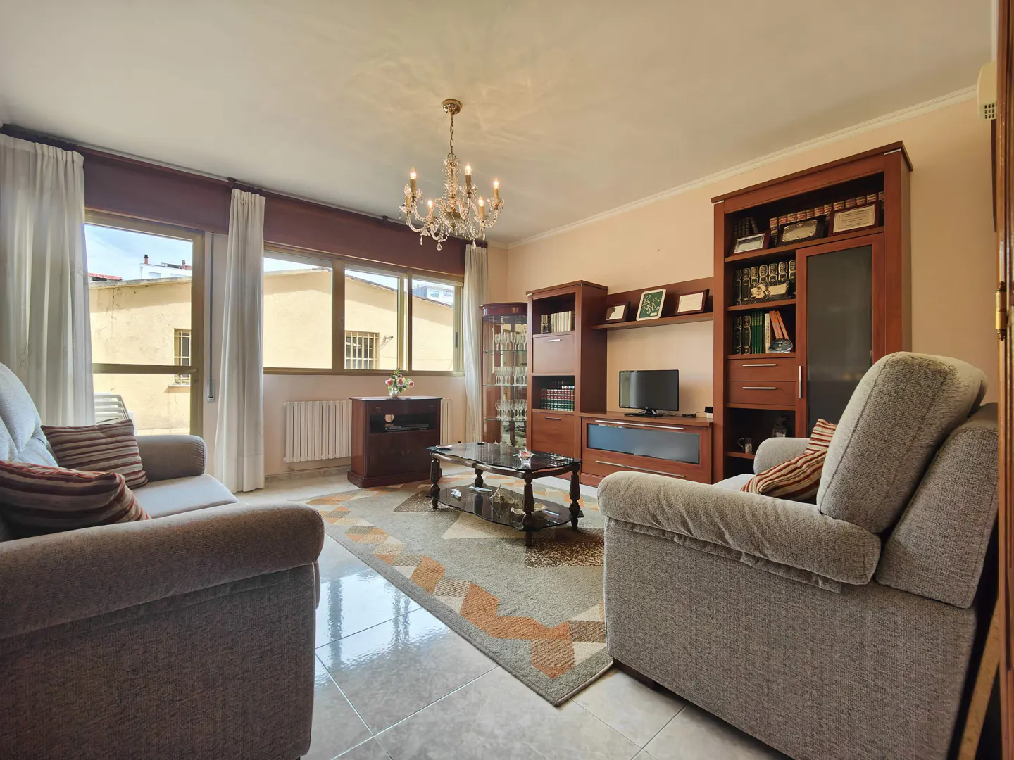 Living room with two grey sofas, a patterned rug, a wooden entertainment unit, and a chandelier under a bright window.