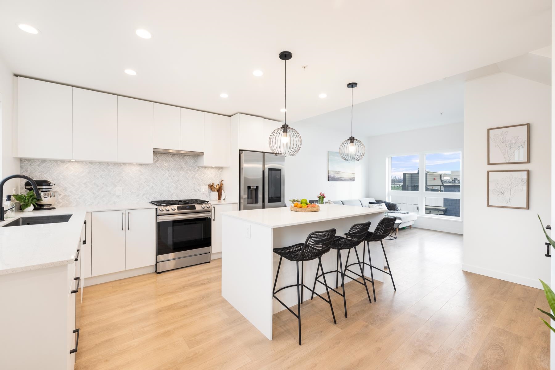 Modern white kitchen with stainless steel appliances, a central island with three black stools, and light wood flooring.